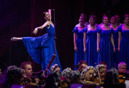 Na pierwszym planie Artystka Baletu we współczesnej, niebieskiej sukience, wykonująca choreografię taneczną. W tle Artyści Chóru również w strojach współczesnych