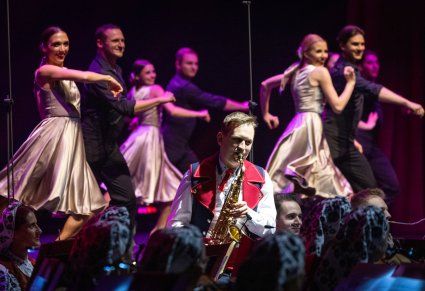 Na pierwszym planie Artysta Orkiestry w kostiumie wilanowskim, grający na saksofonie,  w tle cześć Artystów Baletu w kostiumach współczesnych podczas wykonywania choreografii tanecznej