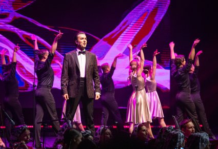 Zbliżenie na Solistę z chóru, za nim część Artystów Baletu w kostiumach współczesnych podczas wykonywania choreografii tanecznej