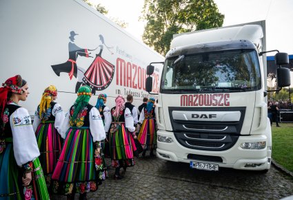 Pary Artystów w kostiumach łowickich przechodzące przy samochodach dostawczych MAZOWSZE