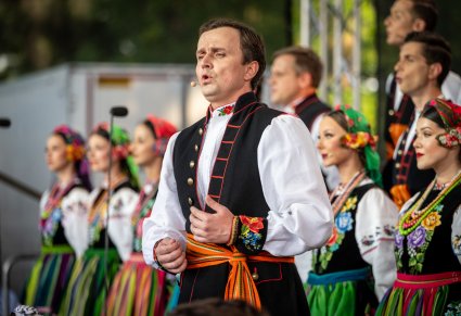 Zbliżenie na Solistę z chóru w kostiumie łowickim. Na drugim planie Artyści z chóru