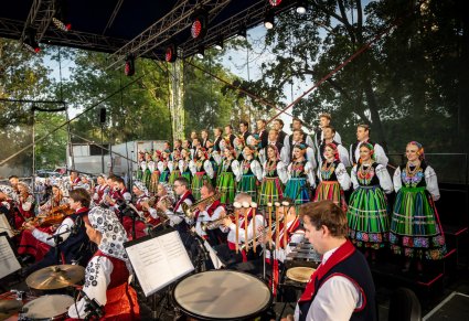 Na pierwszym planie Orkiestra w tle na scenie Artyści z chóru w kostiumach łowickich ustawieni w rzędy
