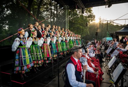 Na scenie Artyści chóru w kostiumach łowickich  ustawieni w rzędy z widoczną na dole Orkiestrą