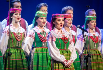 Zbliżenie na śpiewającą uczestniczkę na scenie a w tle chór zespołu "Mazowsze" w kostiumach kurpiowskich