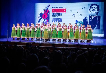 Śpiewająca uczestniczka na scenie a w tle chór zespołu "Mazowsze" w kostiumach kurpiowskich