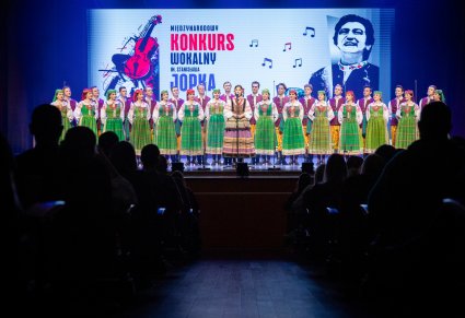 Śpiewająca uczestniczka na scenie a w tle chór zespołu "Mazowsze" w kostiumach kurpiowskich