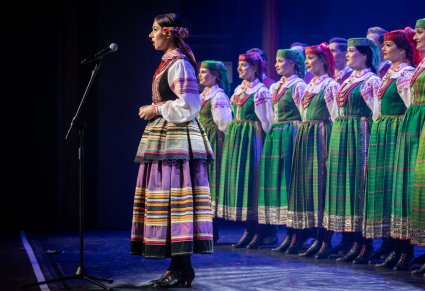 Śpiewająca uczestniczka na scenie a w tle chór zespołu "Mazowsze" w kostiumach kurpiowskich