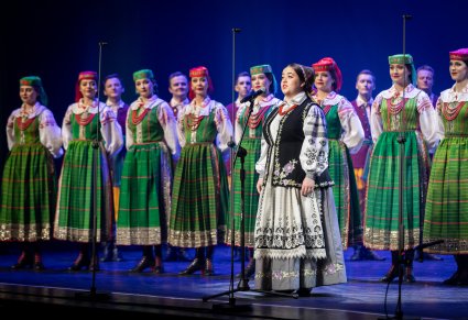 Śpiewająca uczestniczka na scenie a w tle chór zespołu "Mazowsze" w kostiumach kurpiowskich