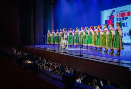 Śpiewająca uczestniczka na scenie a za nią chór zespołu "Mazowsze" w kostiumach kurpiowskich