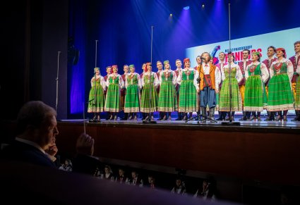 Uczestnik gali a w tle chór zespołu "Mazowsze" w kostiumach kurpiowskich