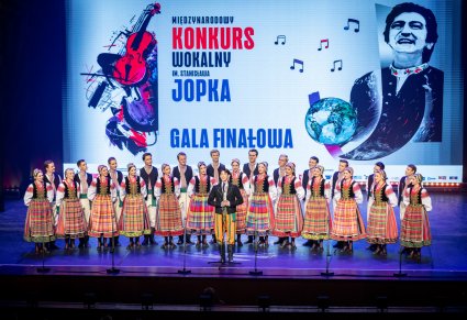 Śpiewający uczestnik gali a w tle chór zespołu "Mazowsze" w kostiumach podlaskich na scenie