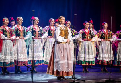 Uczestniczka gali a w tle chór zespołu "Mazowsze" w kostiumach krakowskich
