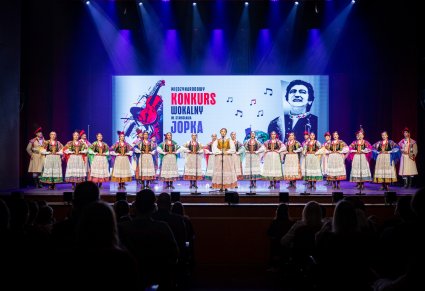 Uczestniczka gali a w tle chór zespołu "Mazowsze" w kostiumach krakowskich