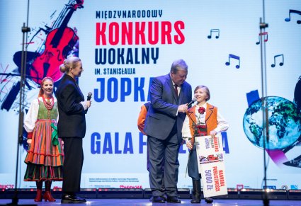 Na scenie Dyrektor Jacek Boniecki wręczający nagrodę dla uczestnika gali obok Anna Maria Jopek wraz z artystą zespołu "Mazowsze" w kostiumie sannickim