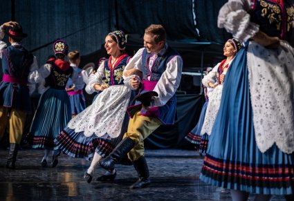 Na pierwszym planie uśmiechnięta para Artystów w kostiumach kaszubskich wykonująca choreografię taneczna na scenie