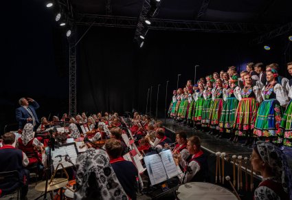 Na scenie Artyści w kostiumach łowickich, niżej orkiestra wraz z Dyrektorem Jackiem Bonieckim, który dyryguje koncert