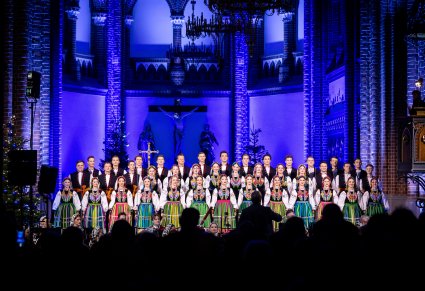 Chór w kostiumach łowickich śpiewa a przed nimi dyrygent i orkiestra