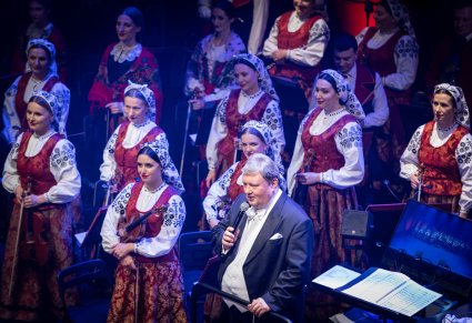 Dyrektor "Mazowsza" przemawia do mikrofonu na tle muzyków z orkiestry w kostiumach wilanowskich.