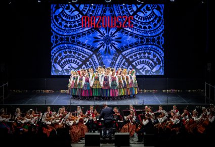 Artyści zespołu Mazowsze w kostiumach opoczyńskich tworzy na scenie koło a przed nimi gra orkiestra