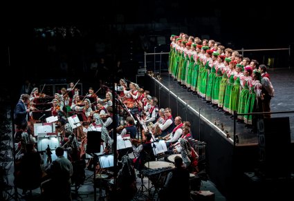 Chór zespołu Mazowsze w kostiumach kurpii białych śpiewa na scenie a przed nimi gra orkiestr