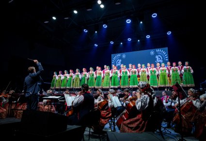 Chór zespołu Mazowsze w kostiumach kurpii białych śpiewa na scenie a przed nimi gra orkiestr