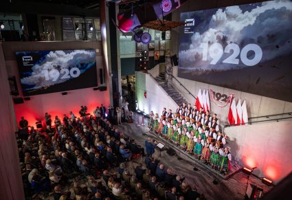 Artyści w czterech rzędach w kostiumach łowickich przed nimi publiczność. Tłem sceny jest napis
