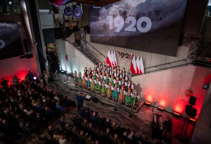 Widok z góry na publiczność i na chór w kostiumach łowickich, na tle sceny jest duży napis 1920