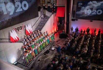 Widok z kolejnego kąta z góry na całe pomieszczenie w którym znajduje się publiczność i Chór w kostiumach łowickich . Nad chórem jest ogromny napis 1920