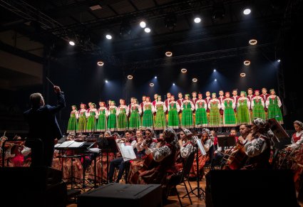Artyści chóru w kostiumach kurpii białych śpiewa na scenie a przed nimi dyrygent i orkiestra