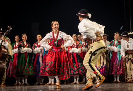 Para tancerzy w kostiumach podhalańskich wykonują taniec góralski na scenie a w tle pozostali artyści