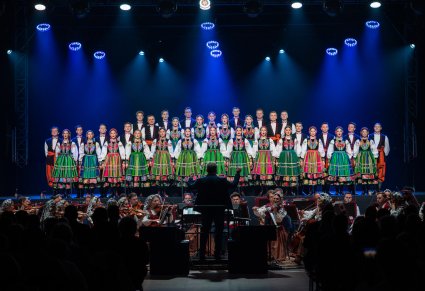 Chór w kostiumach łowickich śpiewa pieśni a przed nimi gra orkiestra