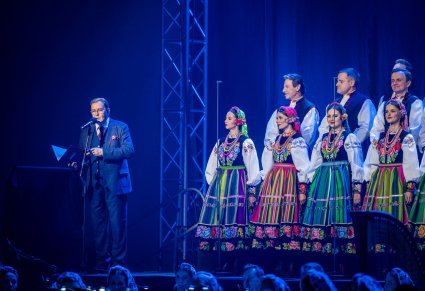 Przemawiająca osoba na scenie, obok widoczny chór