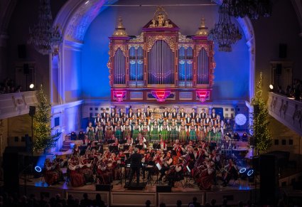 Szerszy widok na chór zespołu Mazowsze, orkiestrę i dyrygenta nagle organów podświetlonych niebieskim światłem