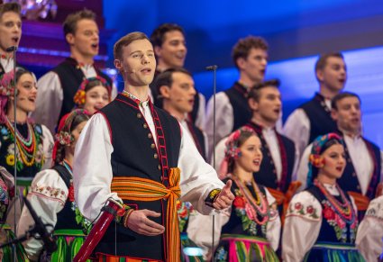 Zbliżenie na śpiewającego solistę a za nim chór w kostiumach łowickich