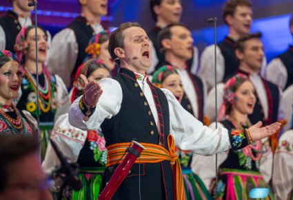 Zbliżenie na śpiewającego solistę a za nim chór w kostiumach łowickich