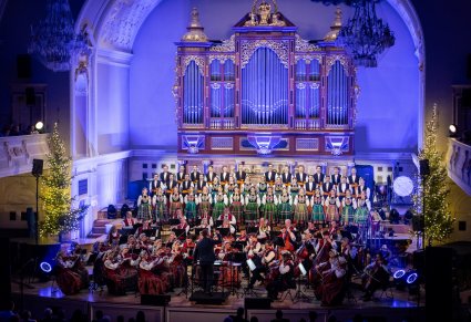 Szerszy widok na chór zespołu Mazowsze, orkiestrę i dyrygenta nagle organów podświetlonych niebieskim światłem