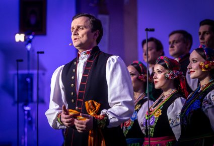 Artysta chóru w kostiumie łowickim podczas wykonywania utworu, w tle widoczny chór