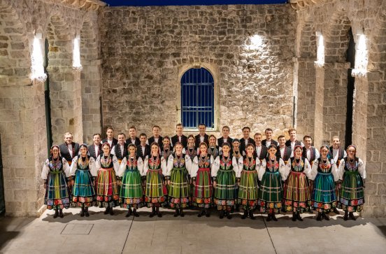  Artyści stojący w dwóch rzędach w kamiennej, oświetlonej sali z łukowymi sklepieniami. Ubrani w kostiumy łowickie  Cała grupa stoi w równym szeregu, uśmiechając się do obiektywu, tworząc harmonijną i barwną kompozycję na tle kamiennej ściany z oknem.