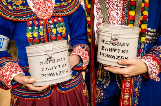  Zbliżenie na puszki "RATUJMY ZABYTKI POWĄZEK" na tle kostiumów podegrodzkich