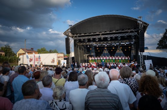  Publiczność przed sceną na której są Artyści w kostiumach kurpiowskich po lewej stronie sceny widać duży samochód z logo MAZOWSZE