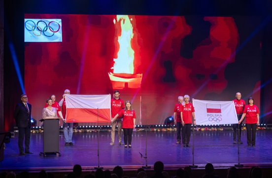  Olimpijczycy na scenie trzymają flagę olimpijską i flagę Polski
