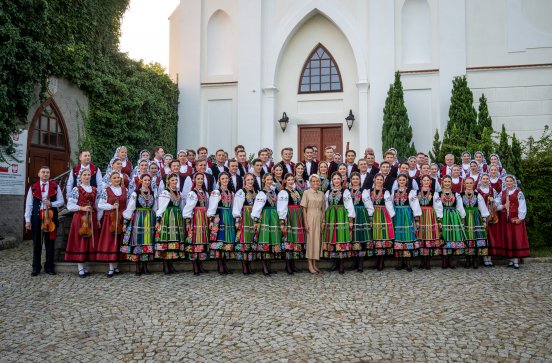  Artyści przed jasnym, zabytkowym budynkiem o wysokich, łukowatych oknach i drzwiach. Większość z nich ma na sobie tradycyjne łowickie stroje ludowe. Kilku artystów trzyma skrzypce i inne instrumenty.