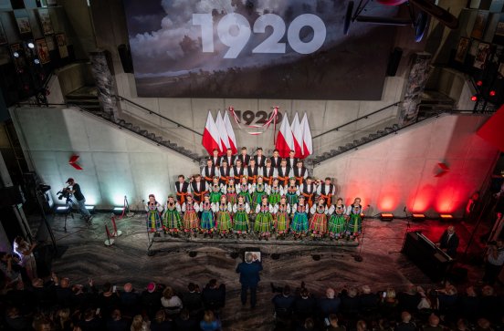  Artyści w kostiumach łowickich ustawieni są na kilku poziomach schodów. Za Artystami zawieszonych jest kilka dużych biało-czerwonych flag, a nad nimi widnieje napis „1920”. Niżej zdjęcia widać Orkiestrę z publicznością